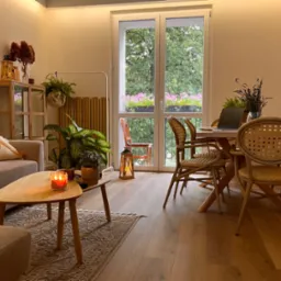 Jasne wnętrze mieszkania z drewnianą podłogą, stołem z krzesłami z rattanu, sofą z poduszką w stylu boho i widokiem na balkon z kwiatami przez okno.