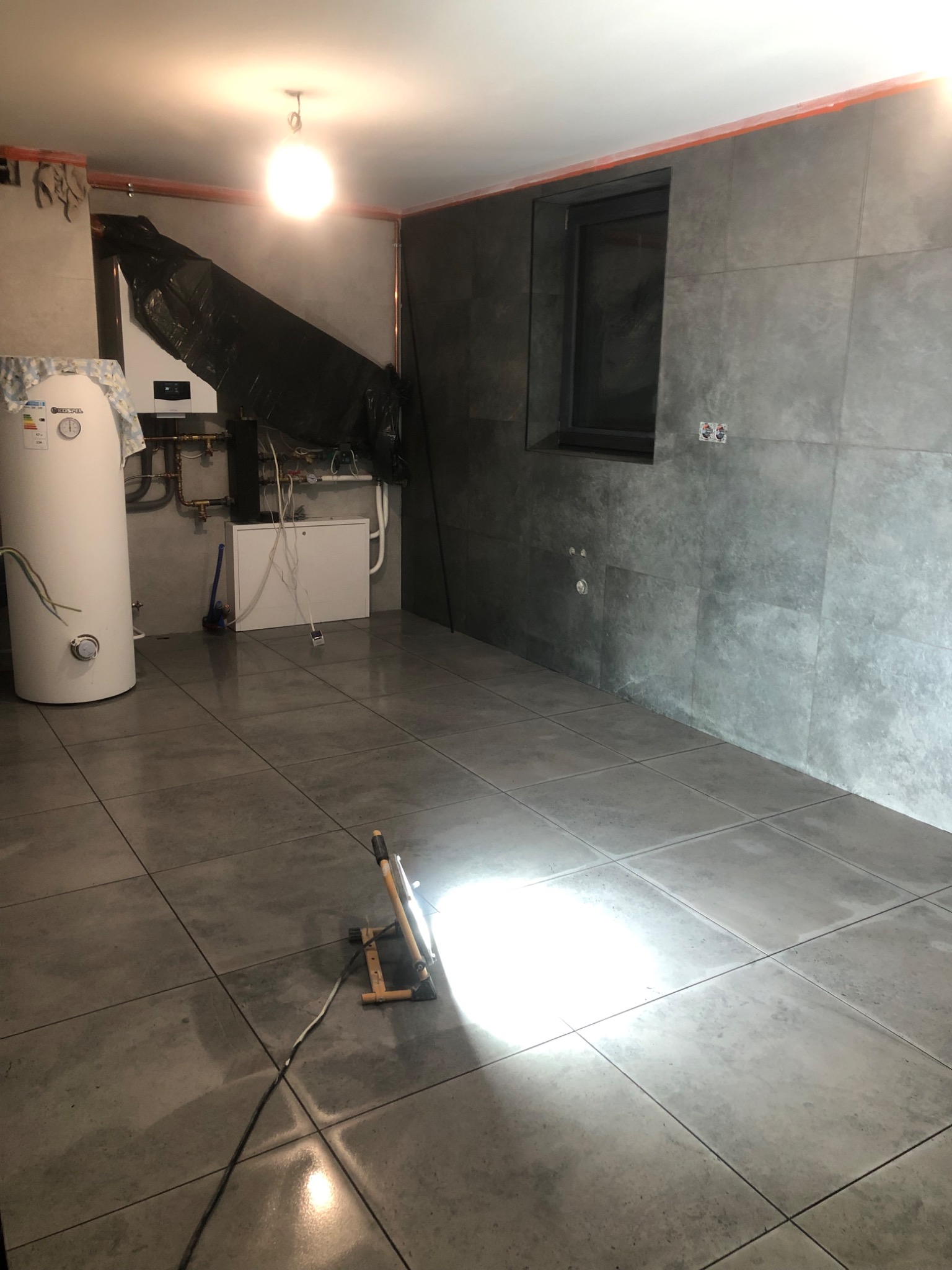 Szare płytki podłogowe i ścienne w surowym pomieszczeniu z kotłem grzewczym, zabezpieczonym folią, oraz przenośną lampą budowlaną na podłodze.