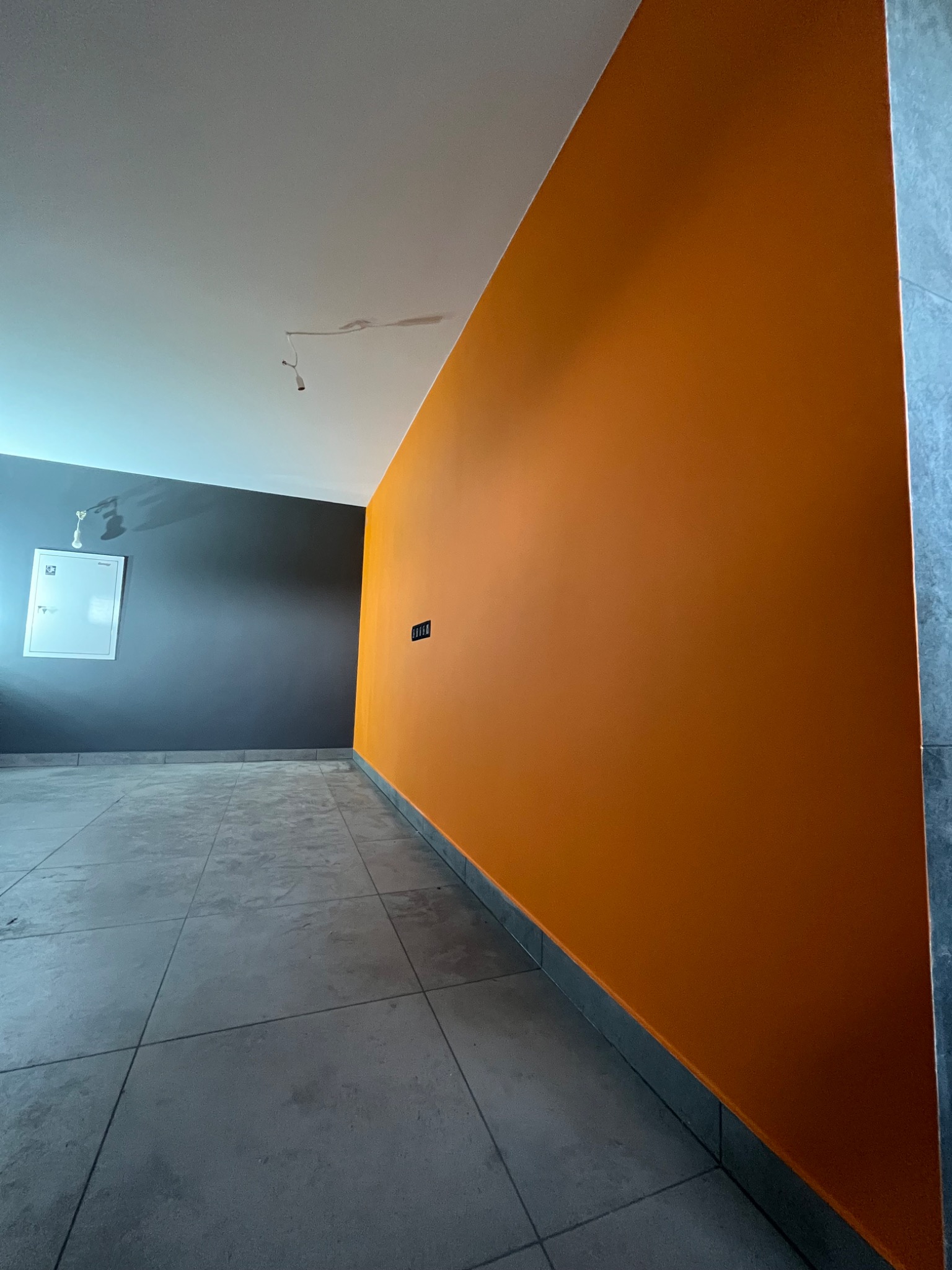 Świeżo pomalowane wnętrze z jedną ścianą w odcieniach pomarańczy, drugą w kolorze grafitowym, szara podłoga z płytek i wiszący kabel od oświetlenia.