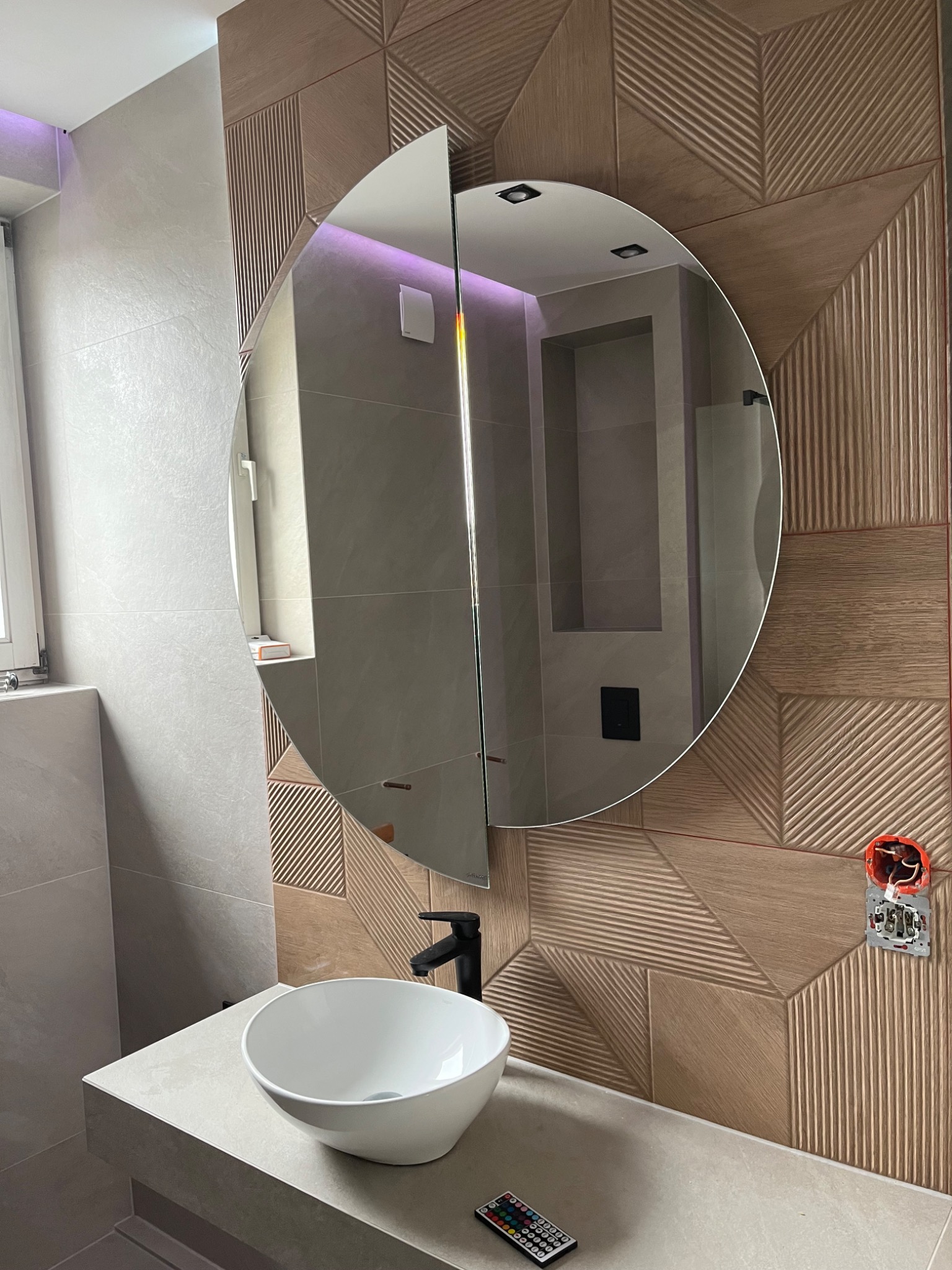 Nowoczesna łazienka z okrągłym lustrem, dekoracyjną ścianą z drewnopodobnych paneli ułożonych w geometryczny wzór, białą umywalką nablatową i czarną armaturą. Widoczna niezakończona instalacja...