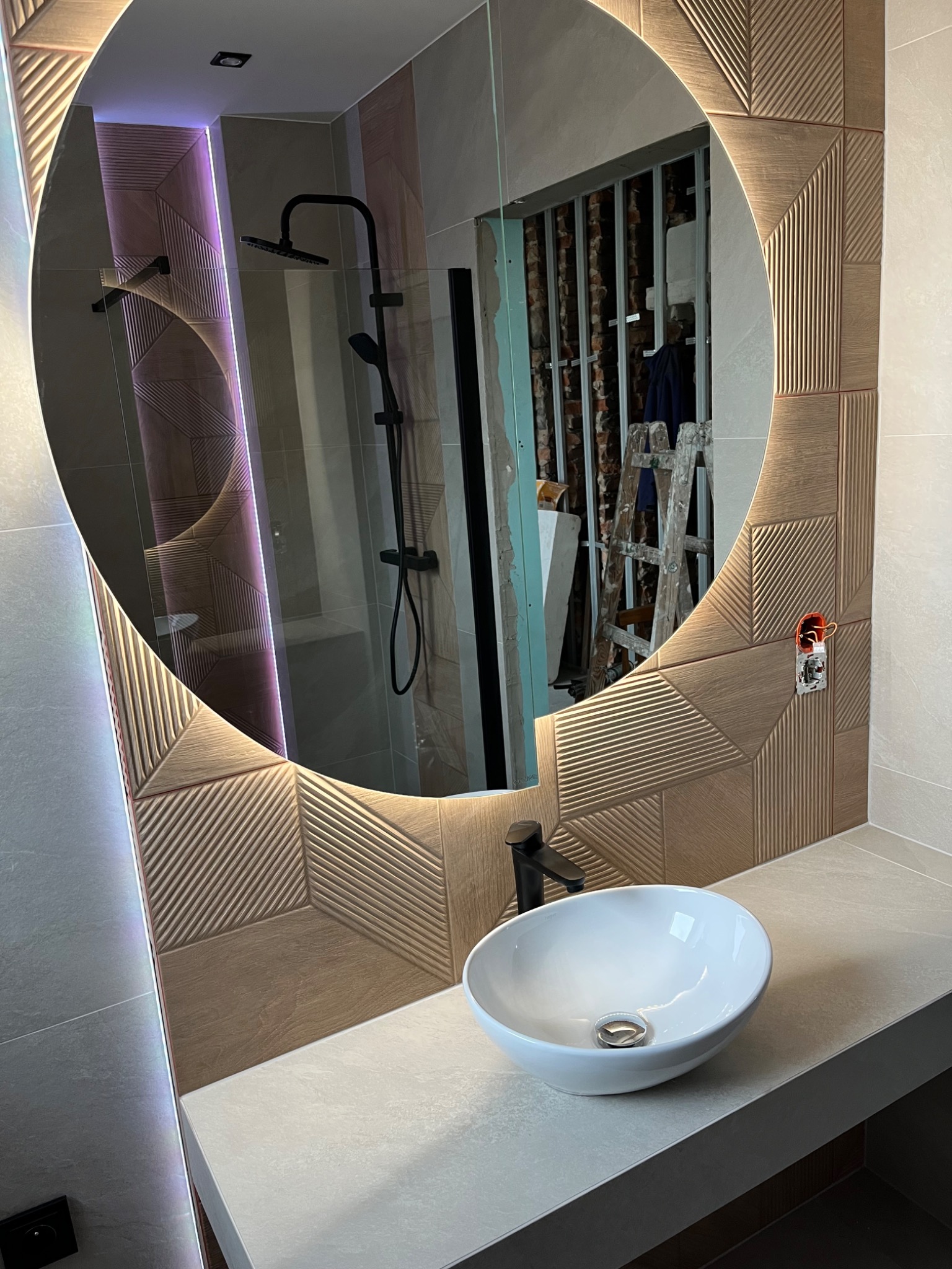 Owalne lustro z podświetleniem LED odbija fragment łazienki w trakcie remontu, widoczna drabina i odsłonięta instalacja elektryczna obok umywalki nablatowej.