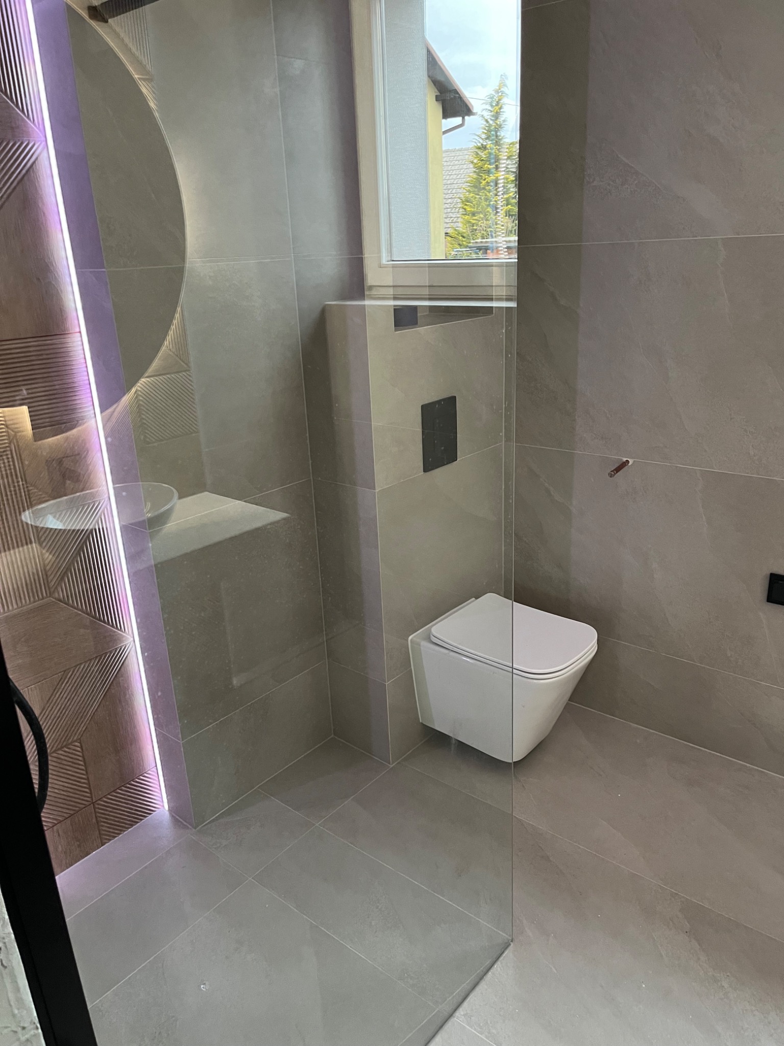 Nowoczesna łazienka z szarymi płytkami imitującymi kamień, podwieszana toaleta, szklana kabina prysznicowa z oświetleniem LED i umywalką na półce.