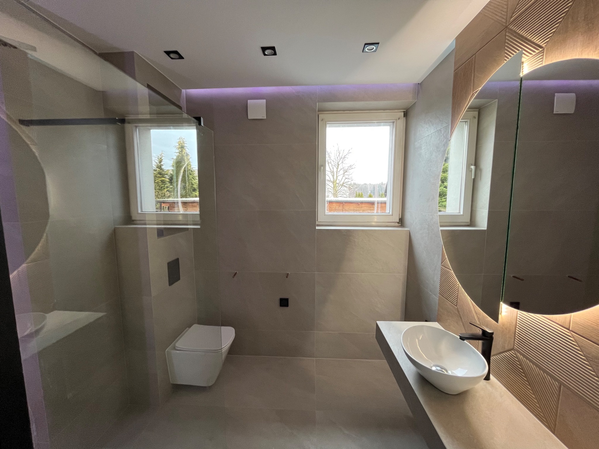 Nowoczesna łazienka w odcieniach szarości z podświetlanym okrągłym lustrem, wiszącą toaletą i umywalką nablatową na betonowym blacie, oświetlenie LED na suficie i widokiem z okna na drzewa.