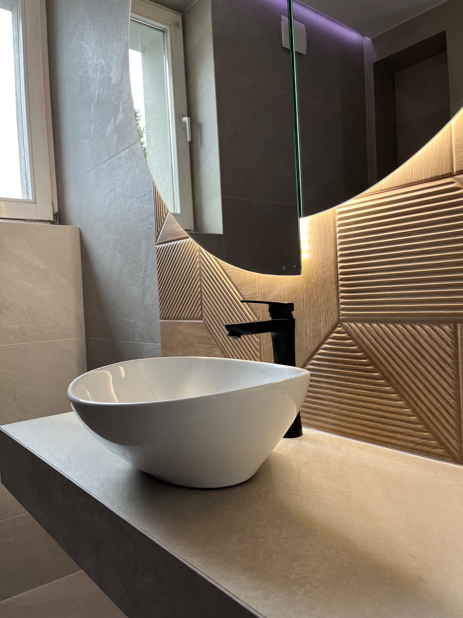 Nowoczesna łazienka z owalną białą umywalką nablatową, czarną baterią i dekoracyjną ścianą z podświetlanym lustrem o geometrycznym wzorze.