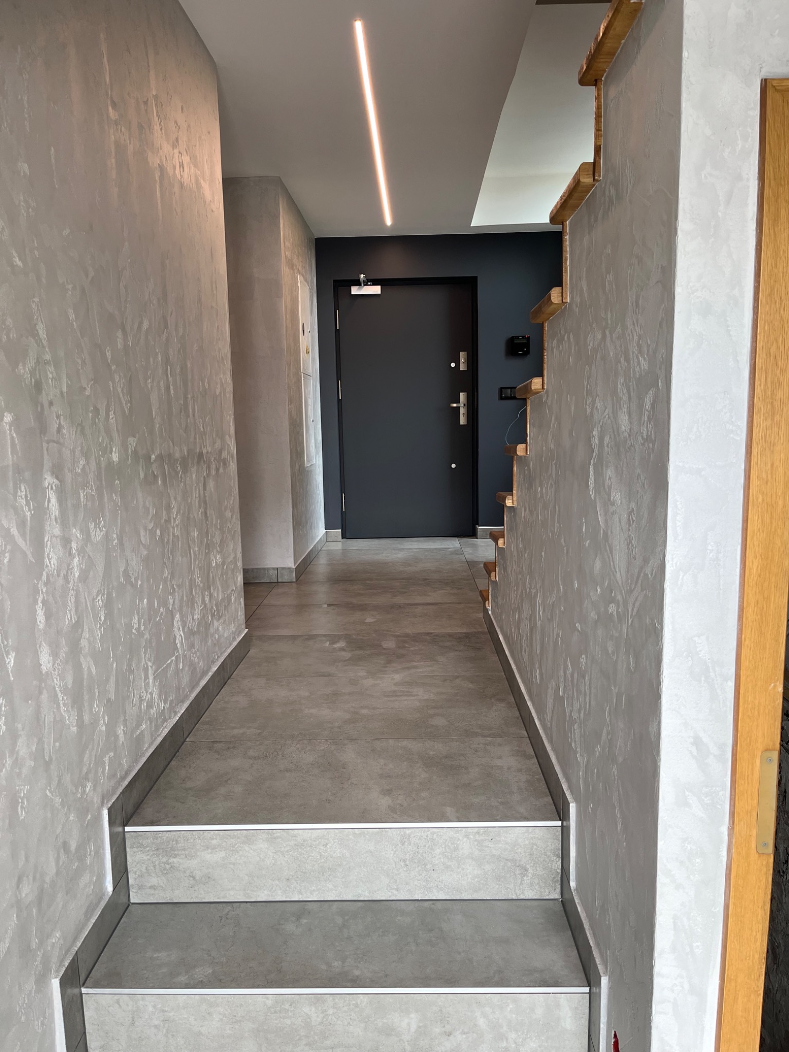 Wnętrze korytarza z szarymi ścianami o fakturze dekoracyjnej, schodami wyłożonymi płytkami w odcieniach szarości z białymi listwami i minimalistycznym oświetleniem LED na suficie, prowadzącego...