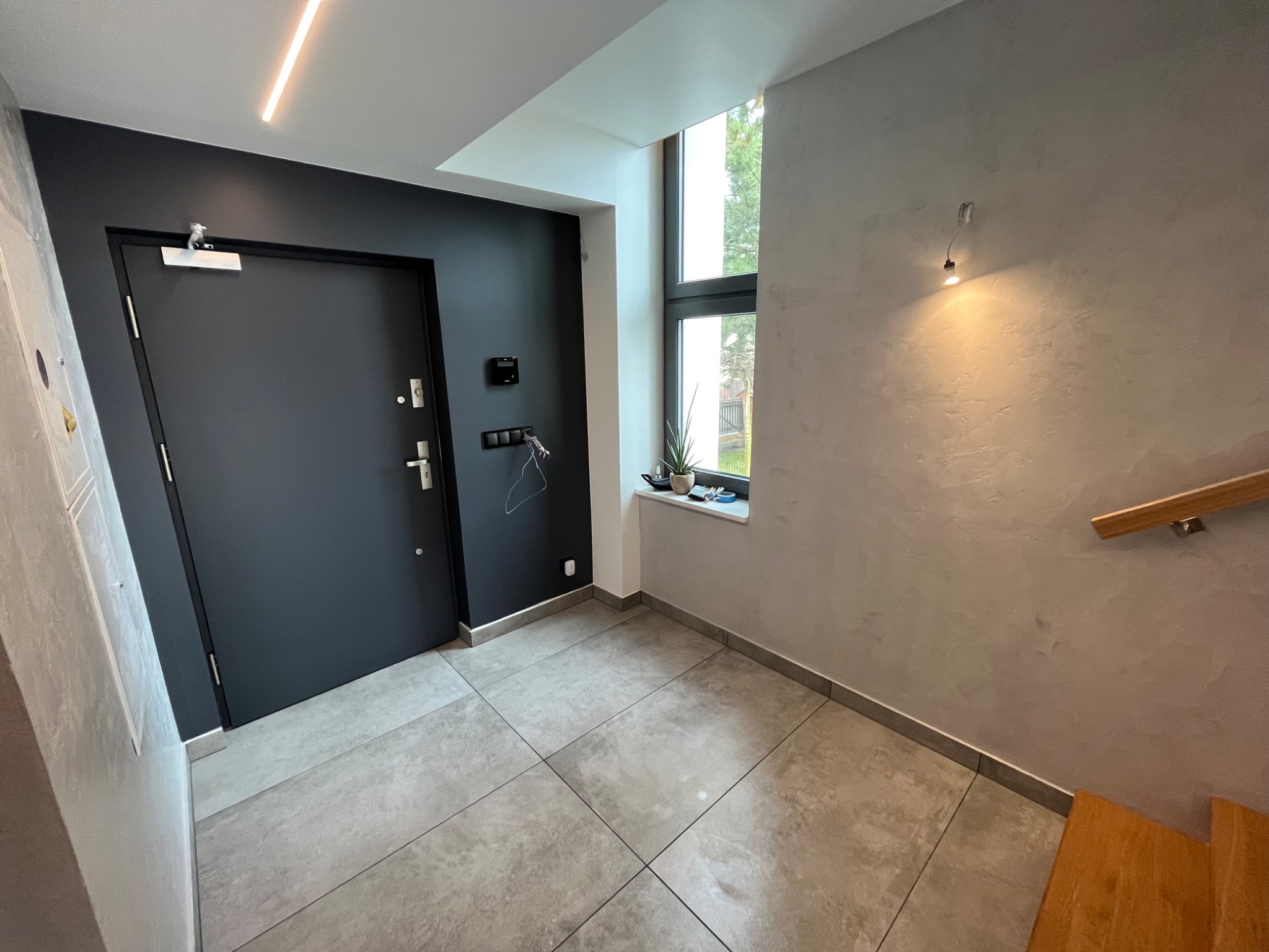 Wnętrze przedpokoju z ciemnymi drzwiami wejściowymi, szarą podłogą z płytek, ścianą z dekoracyjnym tynkiem i oknem z rośliną na parapecie.