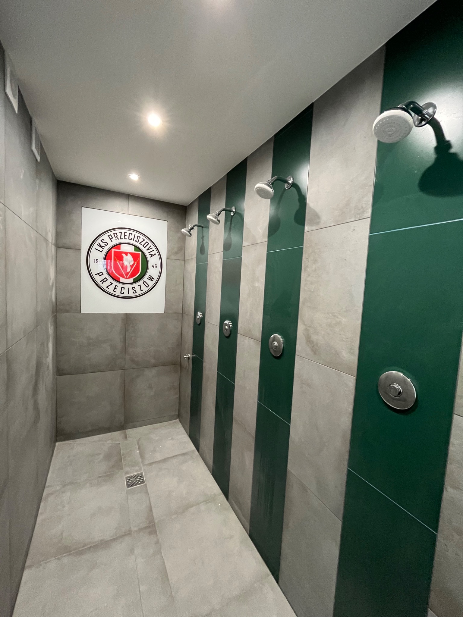 Szare płytki w kabinie prysznicowej z zielonymi pasami i logo klubu sportowego LKS Przeciszovia na ścianie.