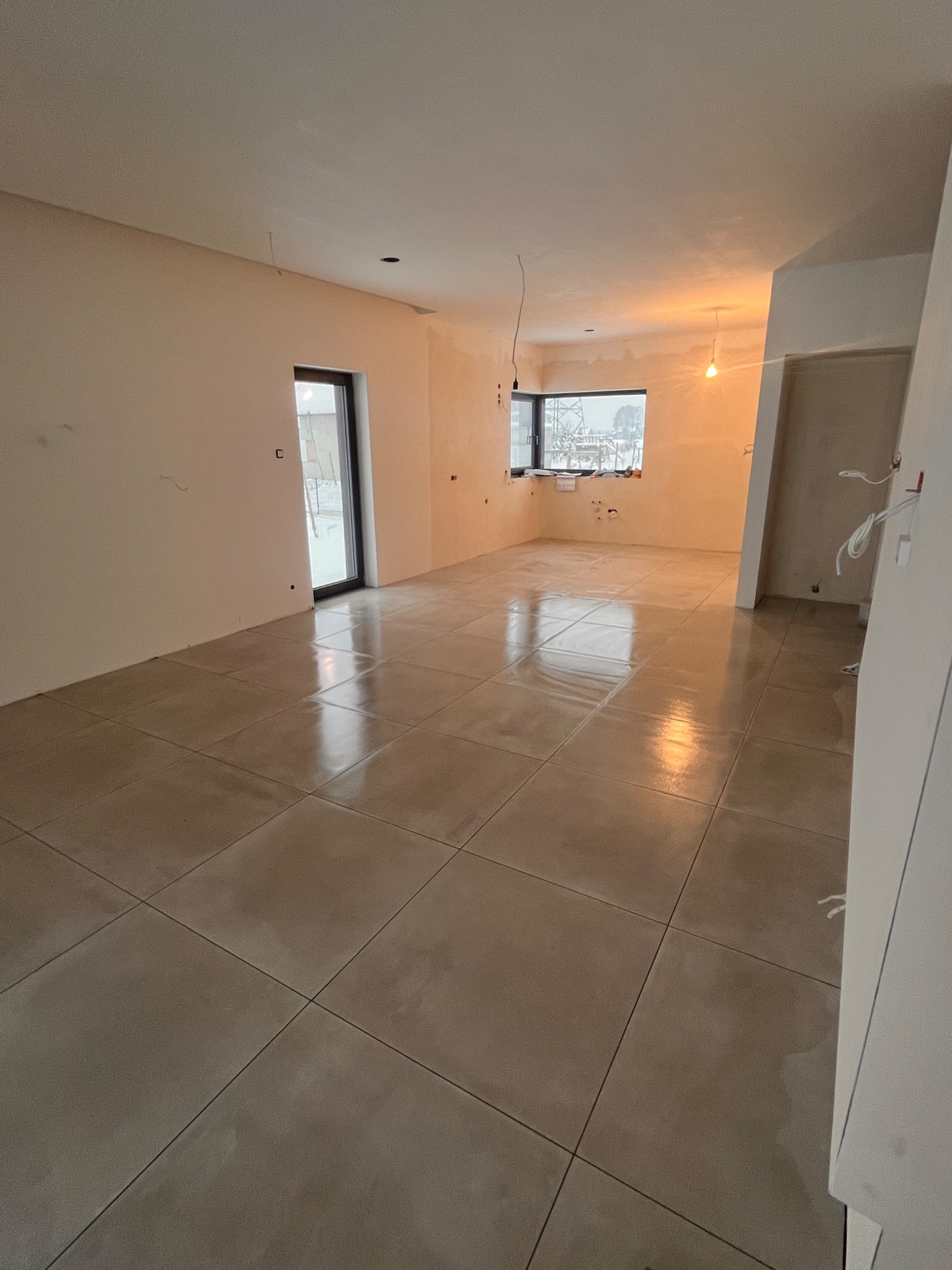 Wnętrze w stanie deweloperskim z ułożoną, błyszczącą, beżową podłogą z płytek, widoczne okno z widokiem na zimowy krajobraz, surowe ściany przygotowane do malowania.