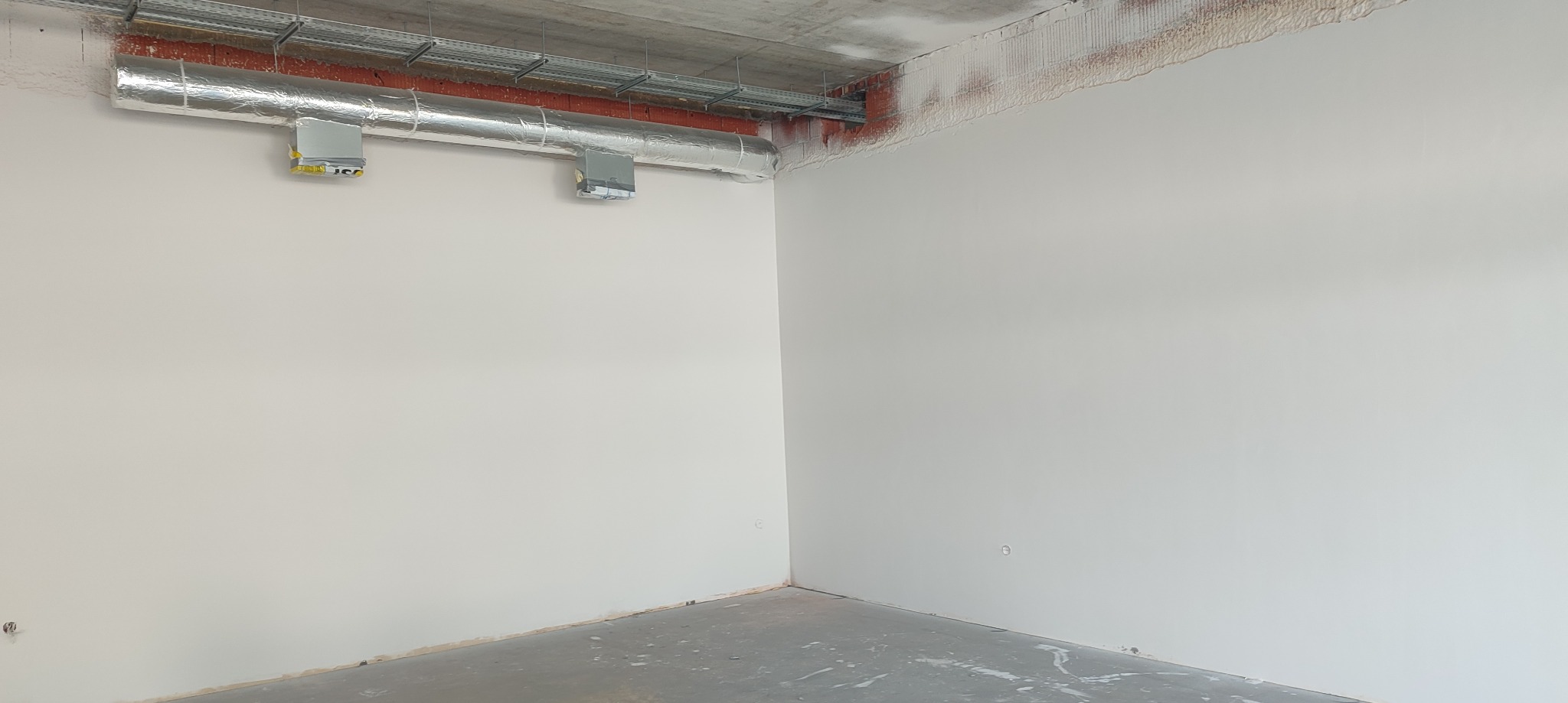 Świeżo pomalowane, jasne ściany w pustym pomieszczeniu z widocznymi instalacjami pod sufitem i surowym betonem. Podłoga betonowa, przygotowana do dalszych prac wykończeniowych.