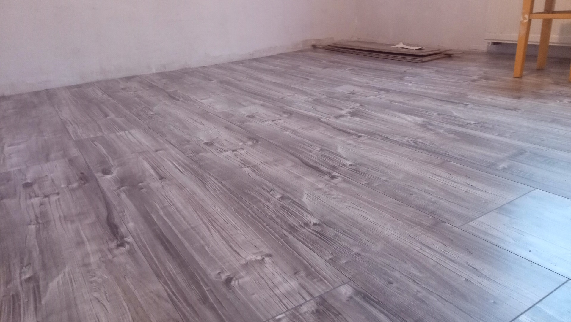Ułożone panele podłogowe o wzorze drewna w odcieniach szarości, widoczny fragment ściany i stos paneli w rogu pomieszczenia.