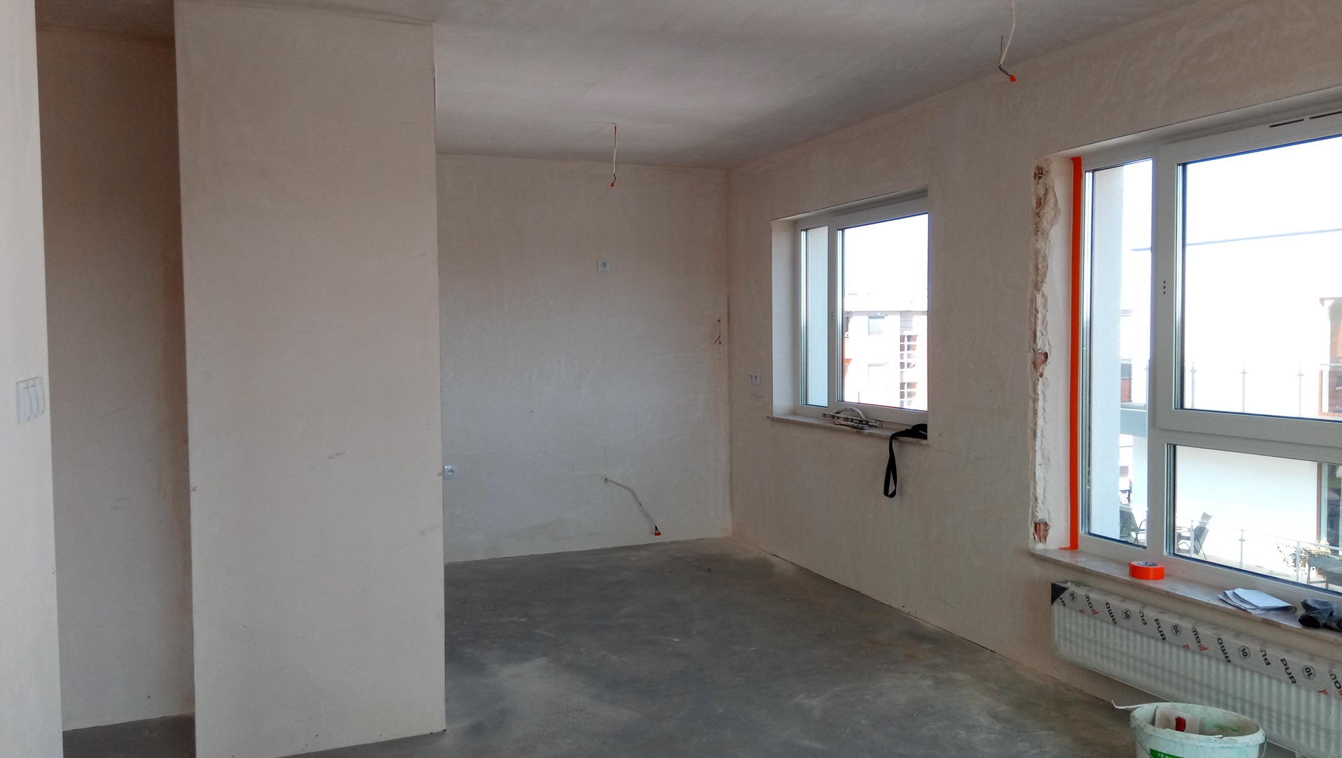 Pokój w stanie deweloperskim z surowymi ścianami po wstępnym wykończeniu, widoczne ślady po instalacjach elektrycznych, okna plastikowe z parapetami, grzejnik z folią ochronną, betonowa podłoga...