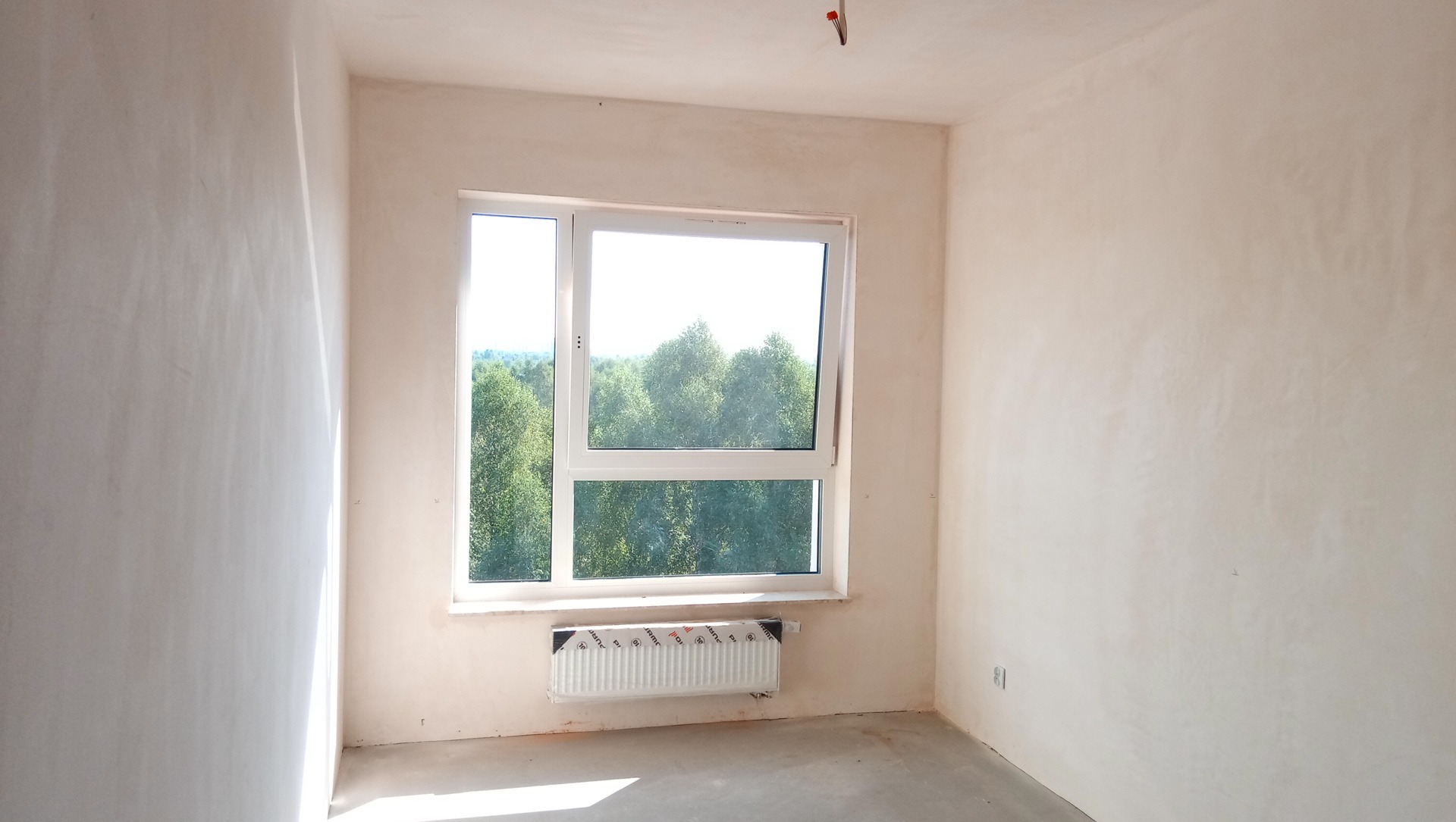 Jasne wnętrze pokoju po wstępnych pracach wykończeniowych ścian, widoczne nierówności podłoża, okno z widokiem na zieleń drzew.
