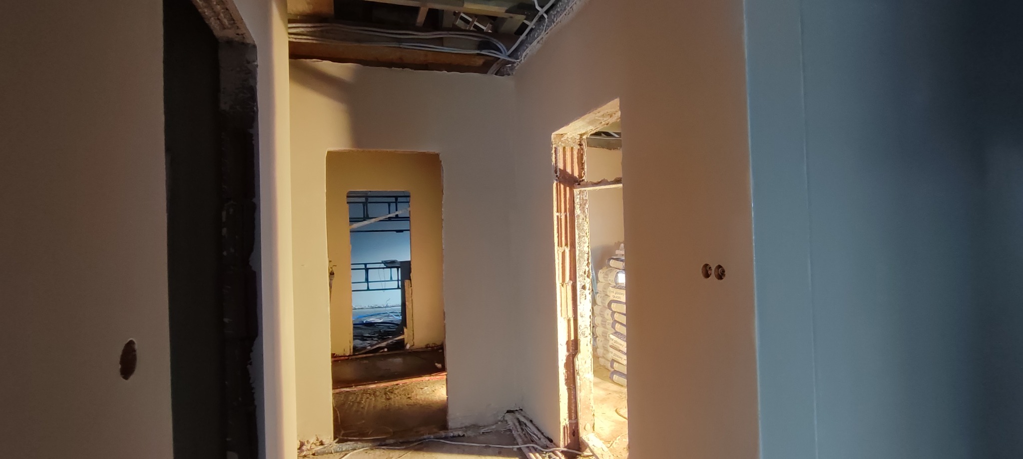Wnętrze w trakcie prac wykończeniowych; ściany pokryte świeżą warstwą wygładzającą, widoczne kable pod sufitem i otwory drzwiowe z surową cegłą; w głębi pomieszczenia worki z materiałem budowlanym.