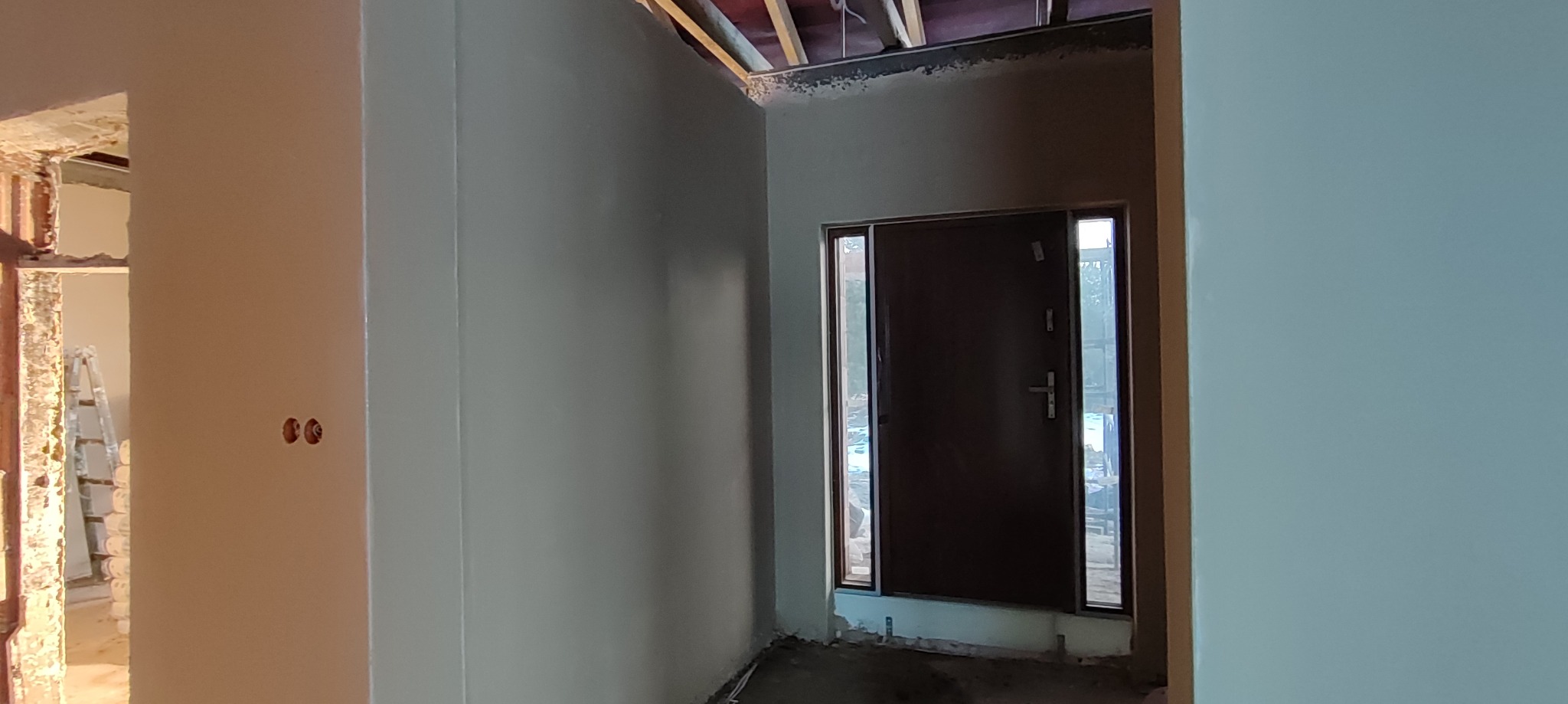 Wnętrze w trakcie remontu: ściany po nałożeniu warstwy wykończeniowej, widoczne gniazdka elektryczne, surowy strop i drzwi wejściowe z ościeżnicą.
