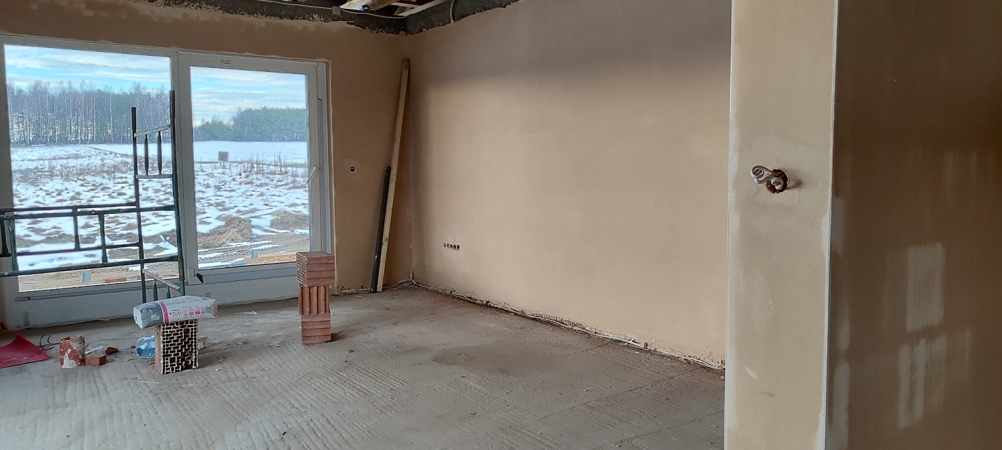 Wnętrze w trakcie prac wykończeniowych: ściany pokryte świeżo nałożoną warstwą, widoczne instalacje elektryczne, okno z widokiem na zimowy krajobraz, cegły i worek materiałów budowlanych...