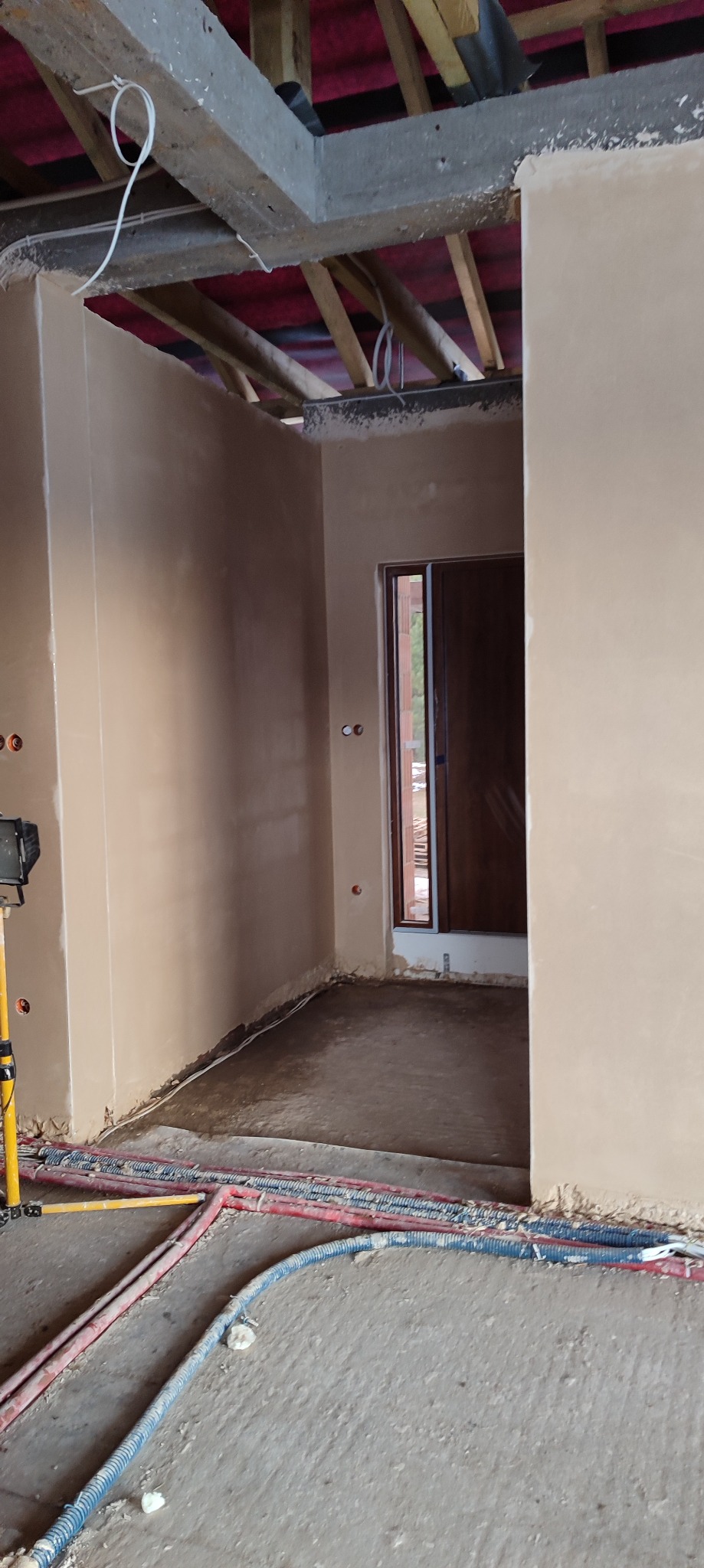Wnętrze w trakcie prac wykończeniowych: ściany pokryte świeżo nałożonym tynkiem, widoczne drewniane elementy konstrukcji dachu, wystające przewody elektryczne i rury instalacyjne na podłodze.