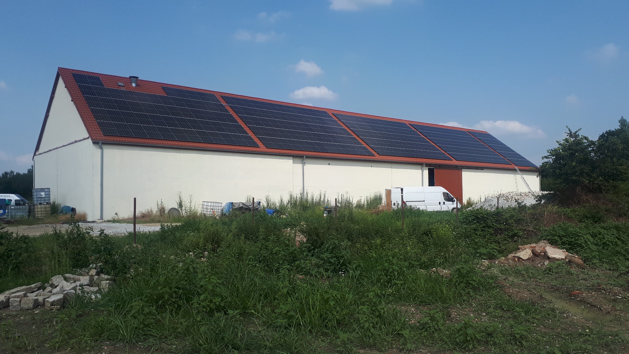 Duży budynek gospodarczy z panelami słonecznymi na czerwonym dachu, biały bus dostawczy zaparkowany obok, teren zarośnięty trawą i chwastami.