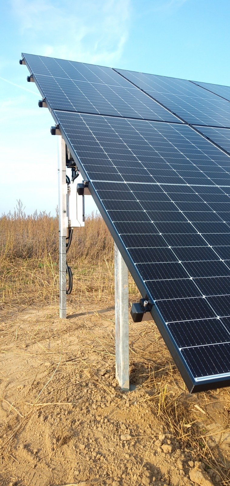 Panele fotowoltaiczne zamontowane na metalowych słupkach w ziemi, widoczne przewody i skrzynka elektryczna, tło z suchą trawą i błękitnym niebem.