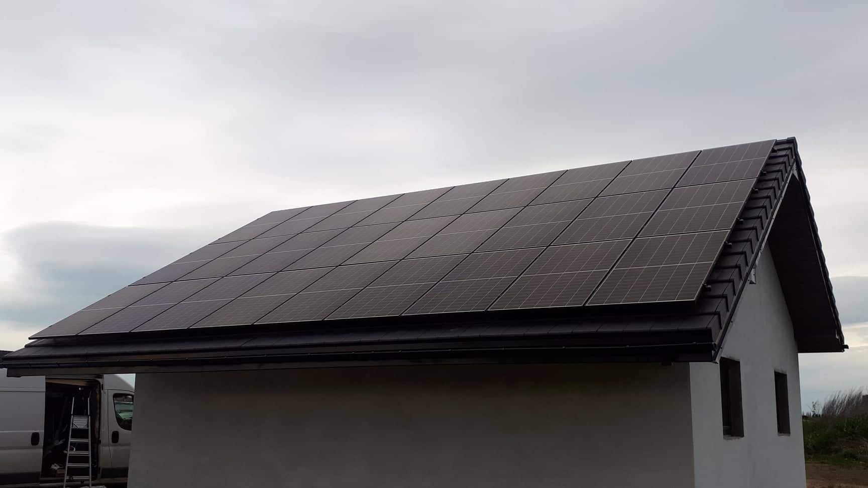 Panele słoneczne na dachu budynku z ciemną dachówką, widoczny fragment białego samochodu dostawczego i drabina.