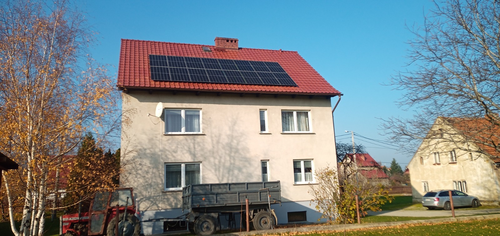 Dwukondygnacyjny dom z czerwonym dachem i panelami słonecznymi, obok drzewa z żółtymi liśćmi, traktor i przyczepa, widok z dołu w słoneczny dzień.
