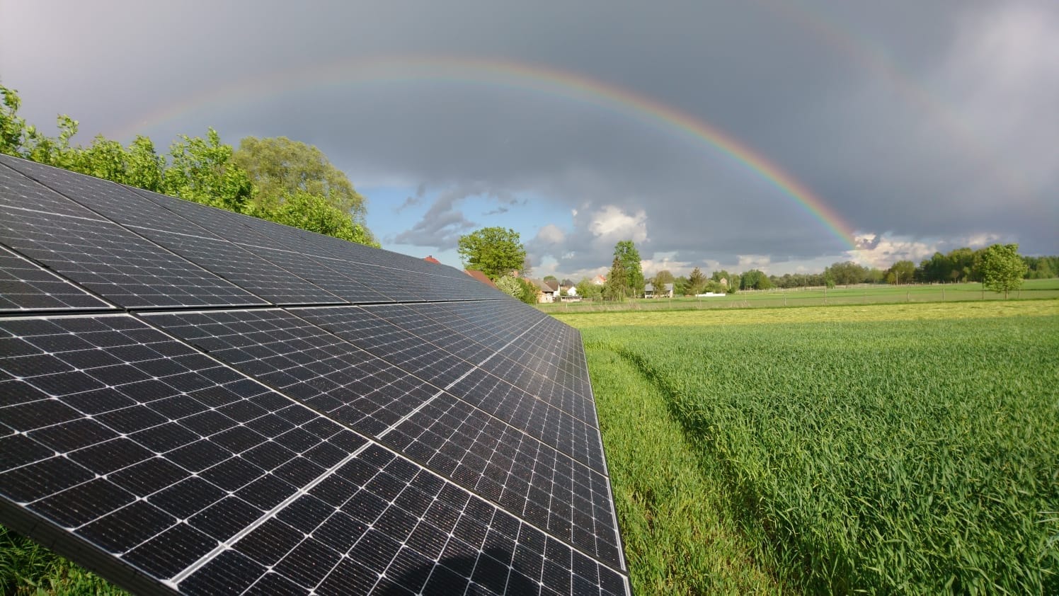 Panel słoneczny na tle zielonego pola i tęczy na pochmurnym niebie, widok z niskiej perspektywy.