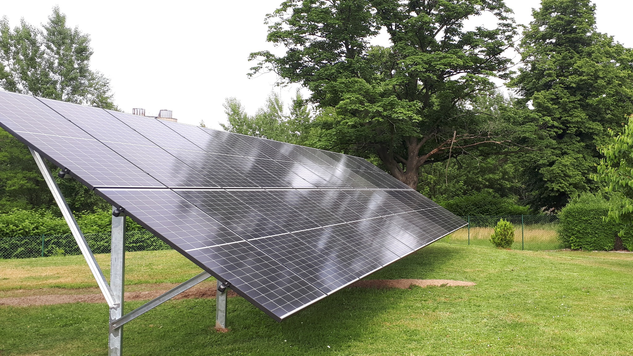 Panel słoneczny na metalowej konstrukcji na trawniku, w tle drzewa i krzewy, widoczne odbicia chmur na panelach.