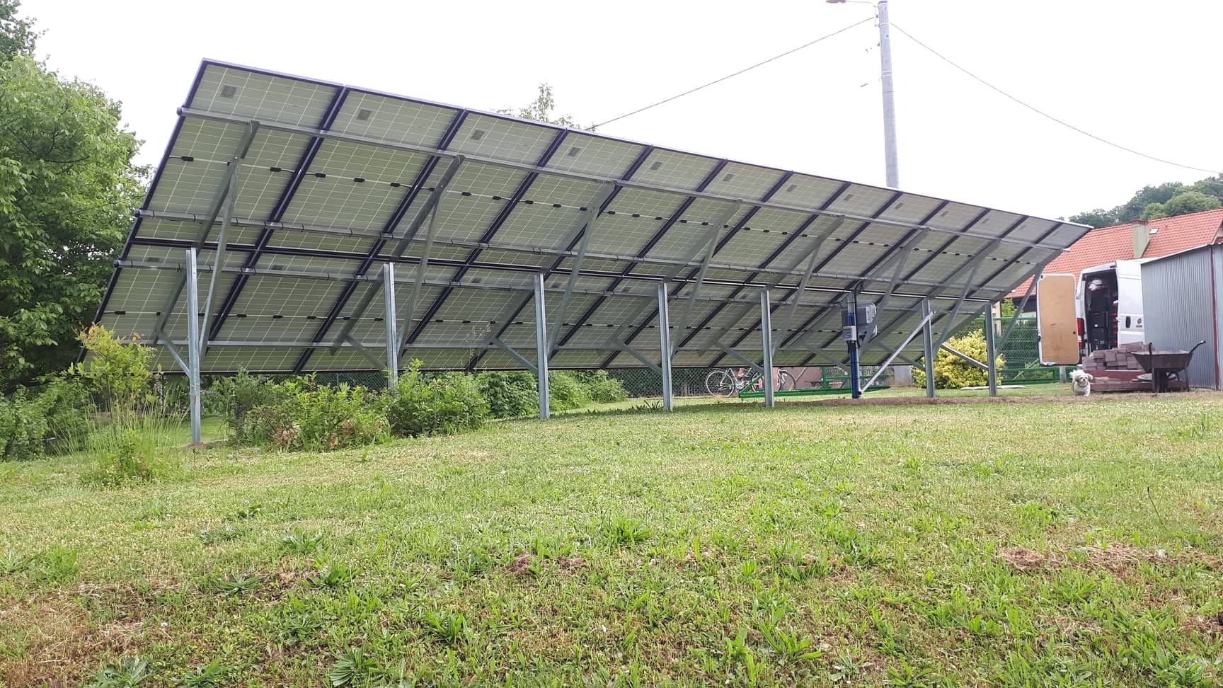 Widok od spodu na panele słoneczne zamontowane na metalowej konstrukcji na trawniku, w tle widoczny rower, budynek gospodarczy i pies.