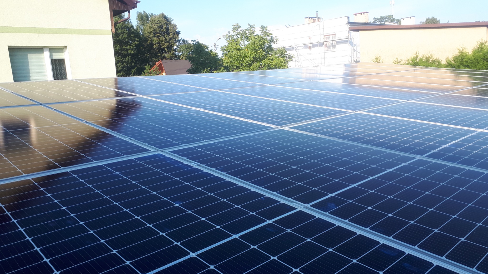 Błyszczące panele słoneczne na dachu budynku w słoneczny dzień, odbijające niebo i otoczenie.