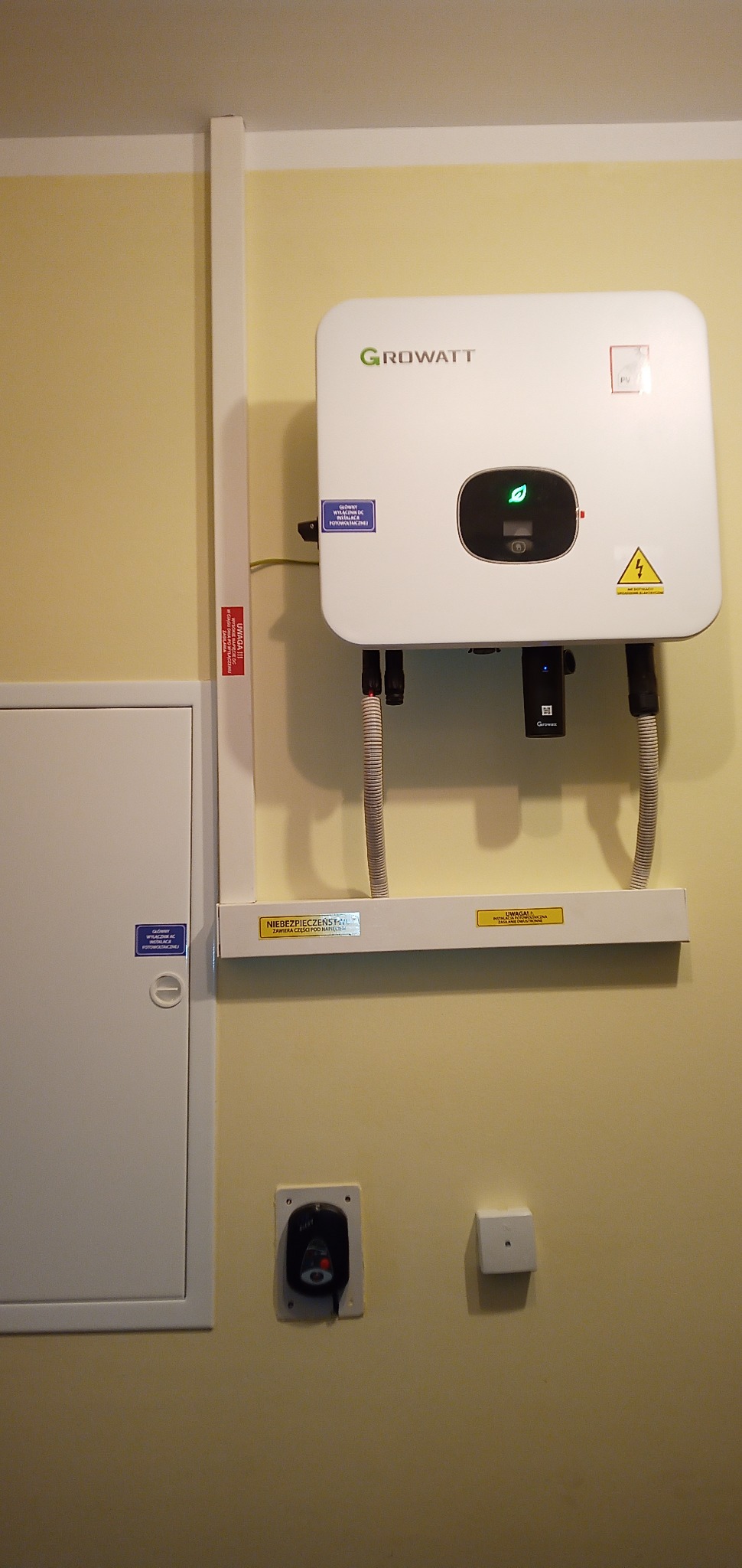 Biały inwerter Growatt zamontowany na ścianie, z widocznymi przewodami, wyłącznikiem i tabliczkami ostrzegawczymi, w pomieszczeniu o żółtej ścianie.
