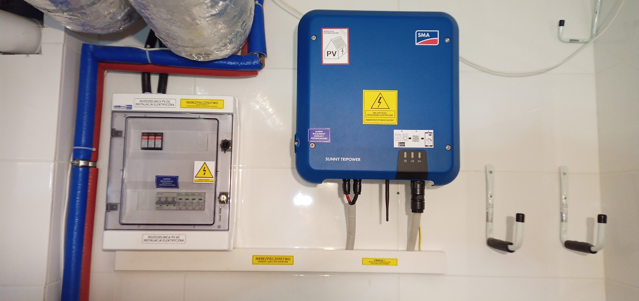 Rozdzielnica PV DC i AC, niebieski inwerter fotowoltaiczny SMA Sunny Tripower, rury instalacyjne w izolacji, zamontowane na białej ścianie wyłożonej kafelkami.