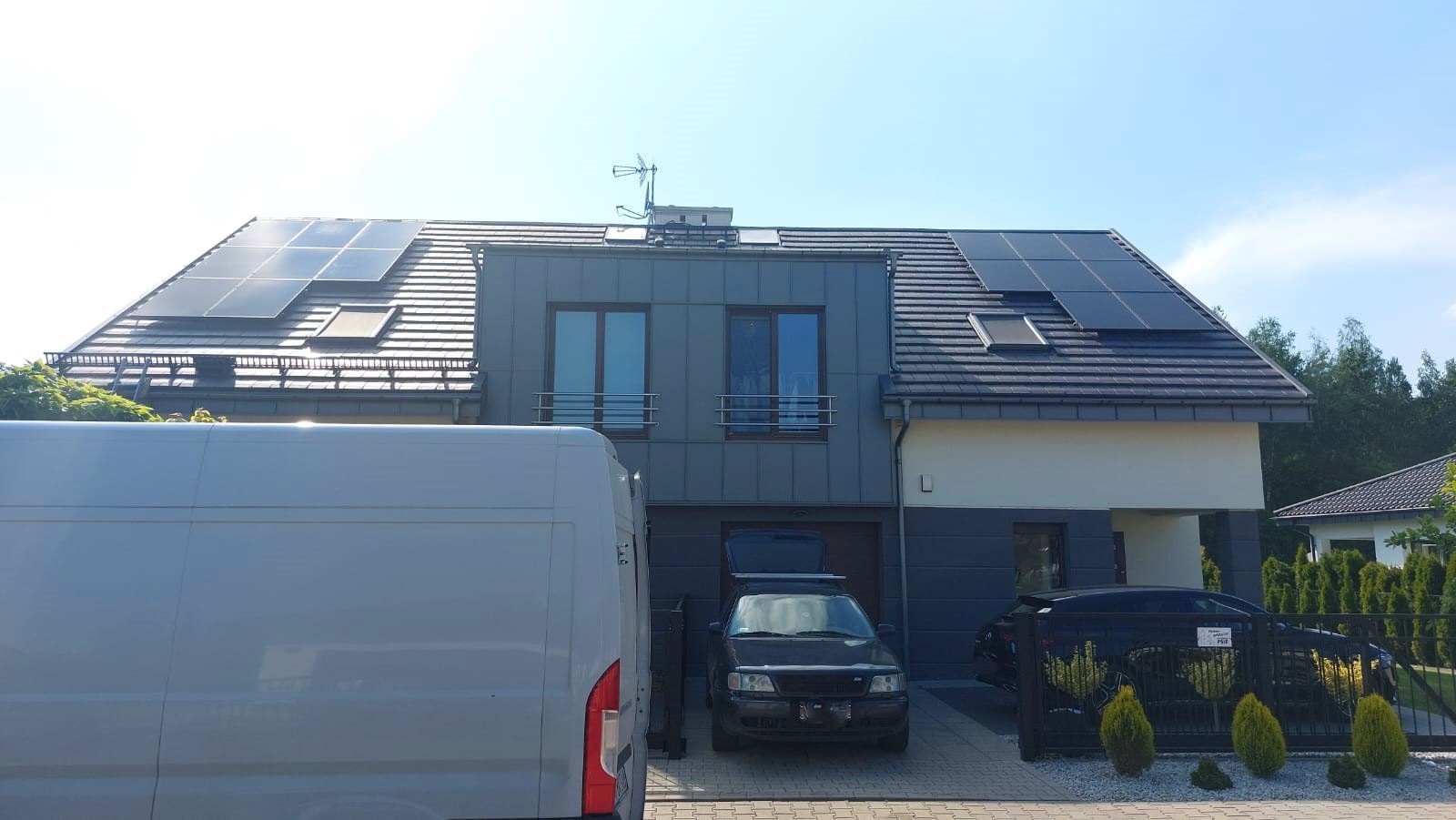 Dom jednorodzinny z panelami słonecznymi na dachu, widoczny biały van i dwa samochody osobowe na podjeździe, brama wjazdowa z tabliczką.