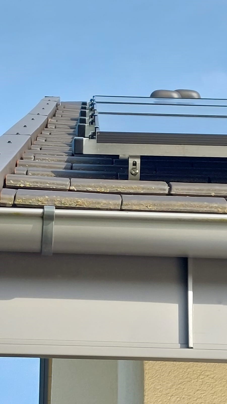 Montaż paneli słonecznych na dachu pokrytym brązową dachówką, widoczne metalowe mocowania i rynna, błękitne niebo w tle.