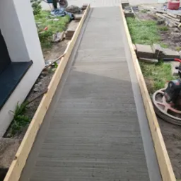 Świeżo wylana, szara posadzka w drewnianej ramie, wygładzona z widocznymi śladami maszyny zacierającej, obok widoczny sprzęt budowlany i taczka.