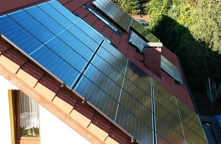 Panele słoneczne na dachu domu z czerwoną dachówką, widoczne okno na poddaszu i komin z cegły.