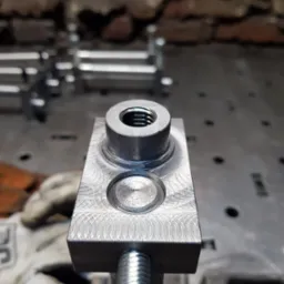 Szlifowany metalowy element z gwintem, zamocowany w uchwycie, widoczne ślady obróbki skrawaniem i rękawice robocze w tle.