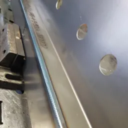 Metalowy element z precyzyjnym spoiną, umieszczony w uchwycie spawalniczym, widoczne otwory w metalu i wygrawerowany napis.