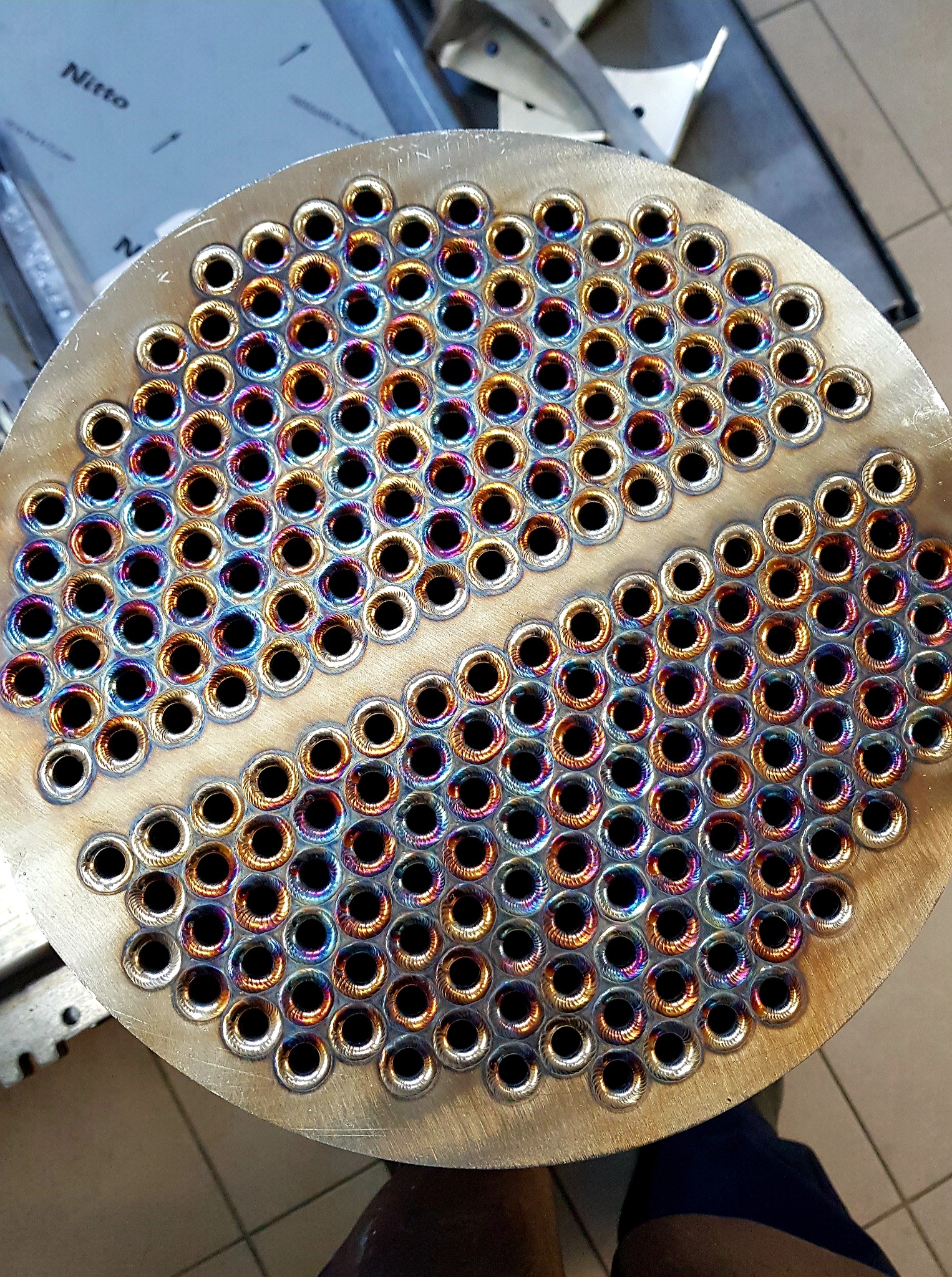 Metalowa płyta z rzędami otworów, każdy z widocznymi śladami spawania, opalizujące kolory na krawędziach otworów, widoczne ślady obróbki na powierzchni płyty.