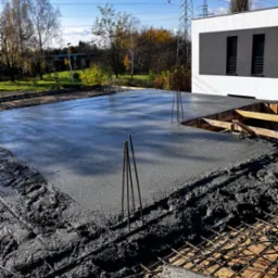 Świeżo wylany, mokry beton na zbrojeniu fundamentu budynku, z widocznymi wystającymi prętami i deskowaniem, w tle nowoczesny budynek z szarymi oknami i słupy wysokiego napięcia pod błękitnym niebem.