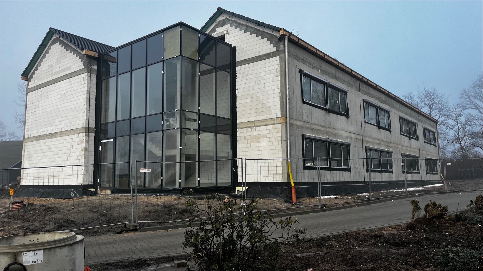 Surowy budynek w trakcie budowy z przeszkloną klatką schodową, widoczne bloczki z betonu komórkowego i tymczasowe ogrodzenie.