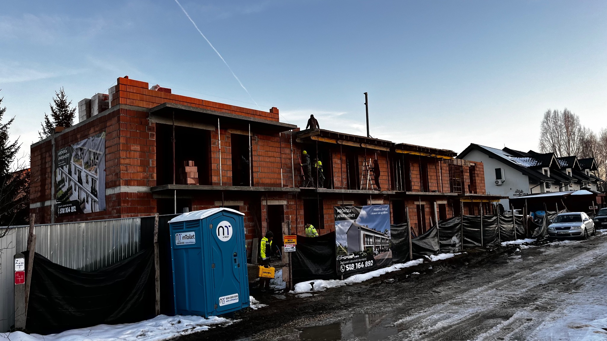 Budowa dwukondygnacyjnego domu murowanego z czerwonej cegły w trakcie realizacji, widoczni robotnicy w żółtych kamizelkach, na pierwszym planie przenośna toaleta i reklama apartamentów.