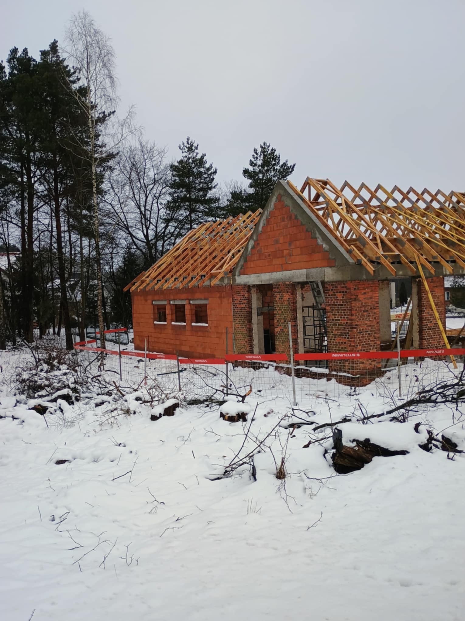 Surowy stan budynku z czerwonej cegły w zimowym krajobrazie, widoczne więźba dachowa i zabezpieczenia budowy.