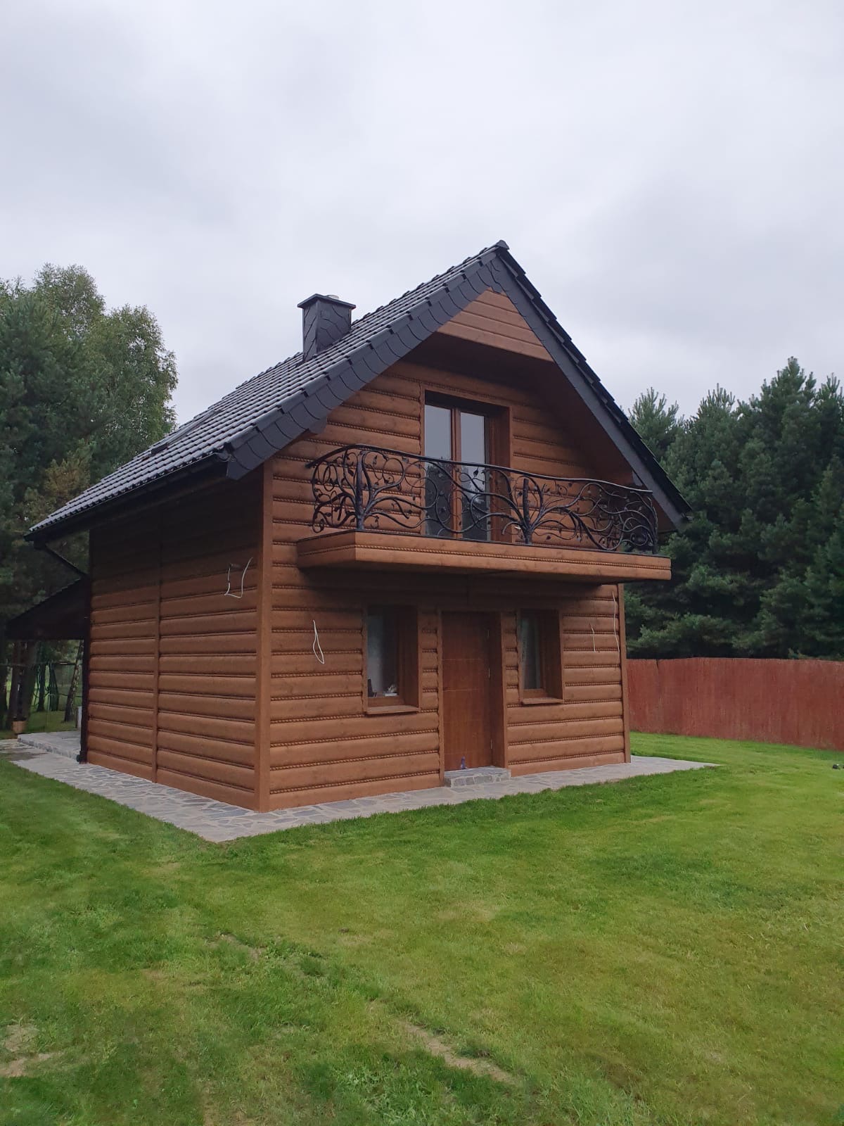 Dwukondygnacyjny dom z bali z czarnym dachem i ozdobnym balkonem z kutego żelaza, otoczony trawnikiem i drzewami.