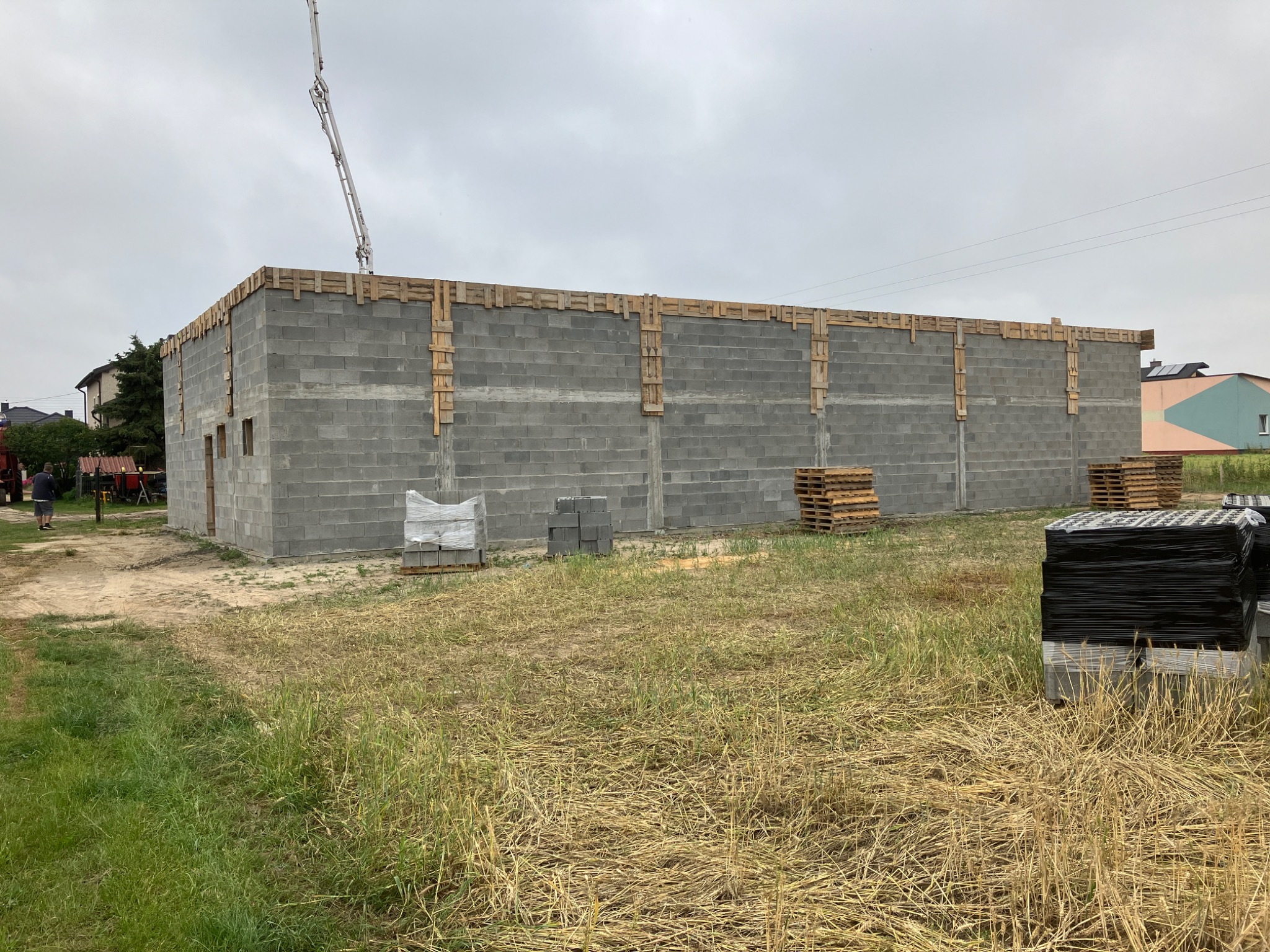 Wznoszenie ścian budynku z bloczków betonowych, z widocznym szalunkiem na górze i pompą do betonu w tle, teren budowy z paletami i materiałami budowlanymi.