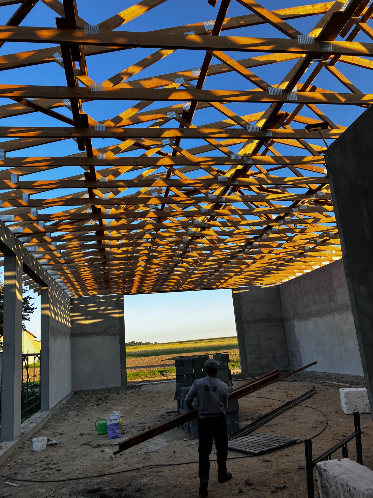 Konstrukcja drewniana dachu w trakcie budowy, widok z wnętrza budynku z betonowymi ścianami, pracownik przenosi elementy konstrukcyjne na tle krajobrazu.