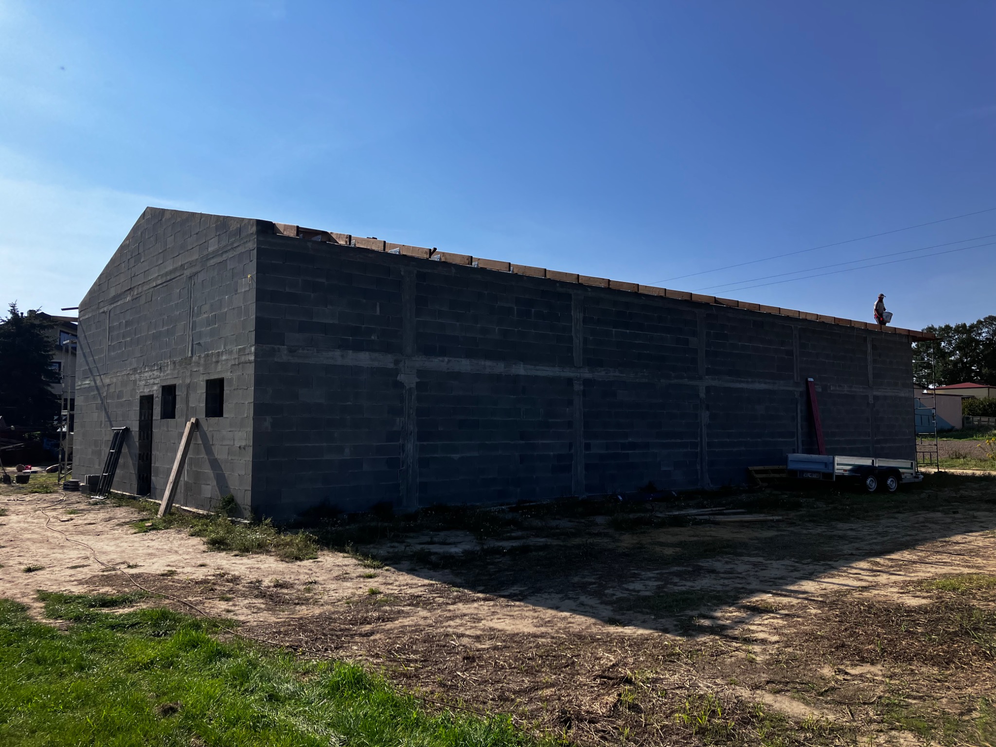 Wznoszenie ścian hali z bloczków betonowych, widoczna przyczepka samochodowa i pracownik na dachu w oddali.