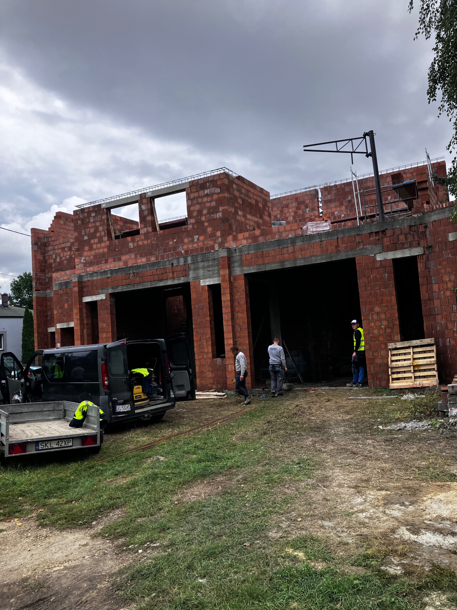 Wznoszenie dwukondygnacyjnego budynku z czerwonej cegły, widoczni pracownicy w kamizelkach odblaskowych, sprzęt budowlany w busie z otwartymi drzwiami, pochmurne niebo.