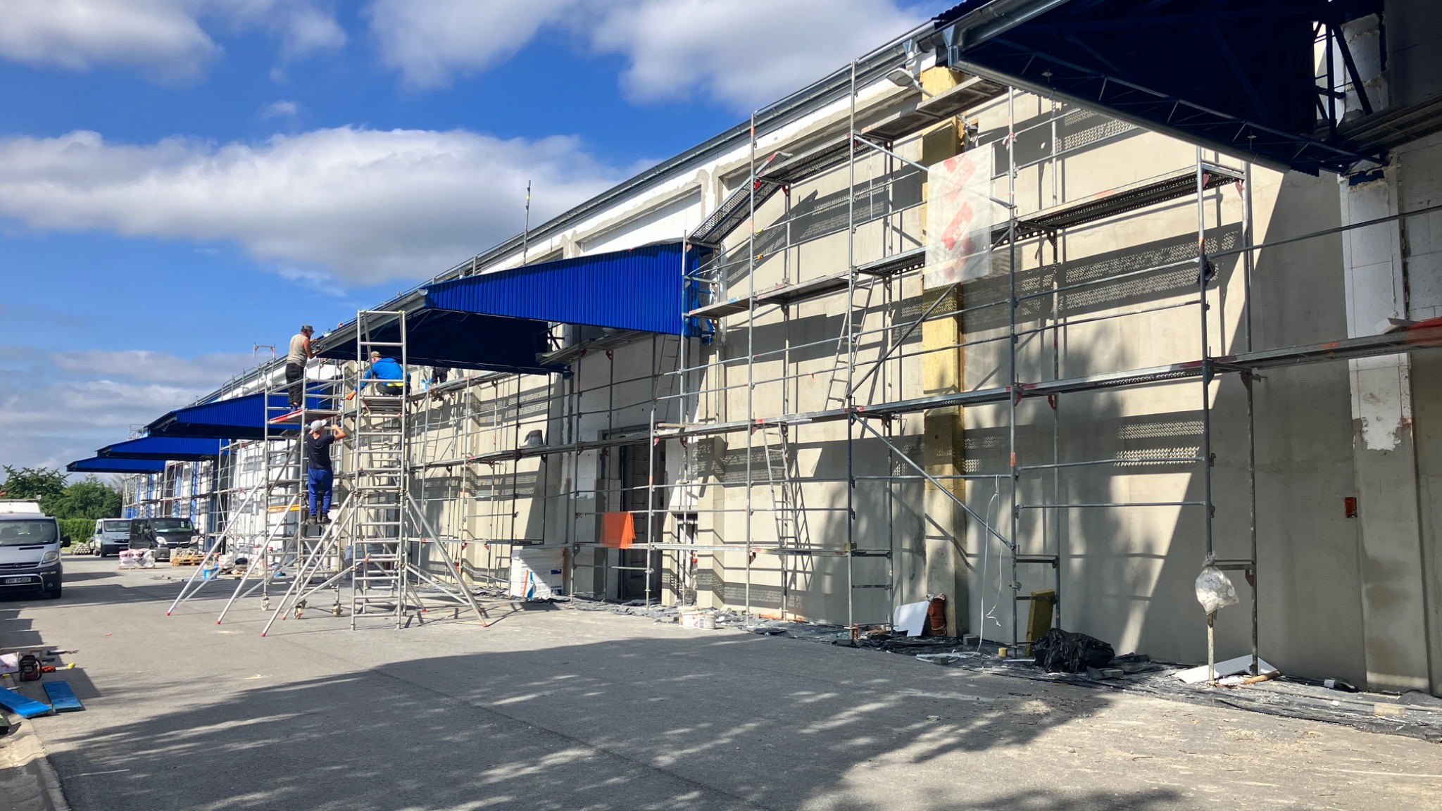 Montaż izolacji na ścianie hali przemysłowej z użyciem rusztowań, widoczne elementy konstrukcji stalowej i obróbki blacharskiej nad wjazdem.