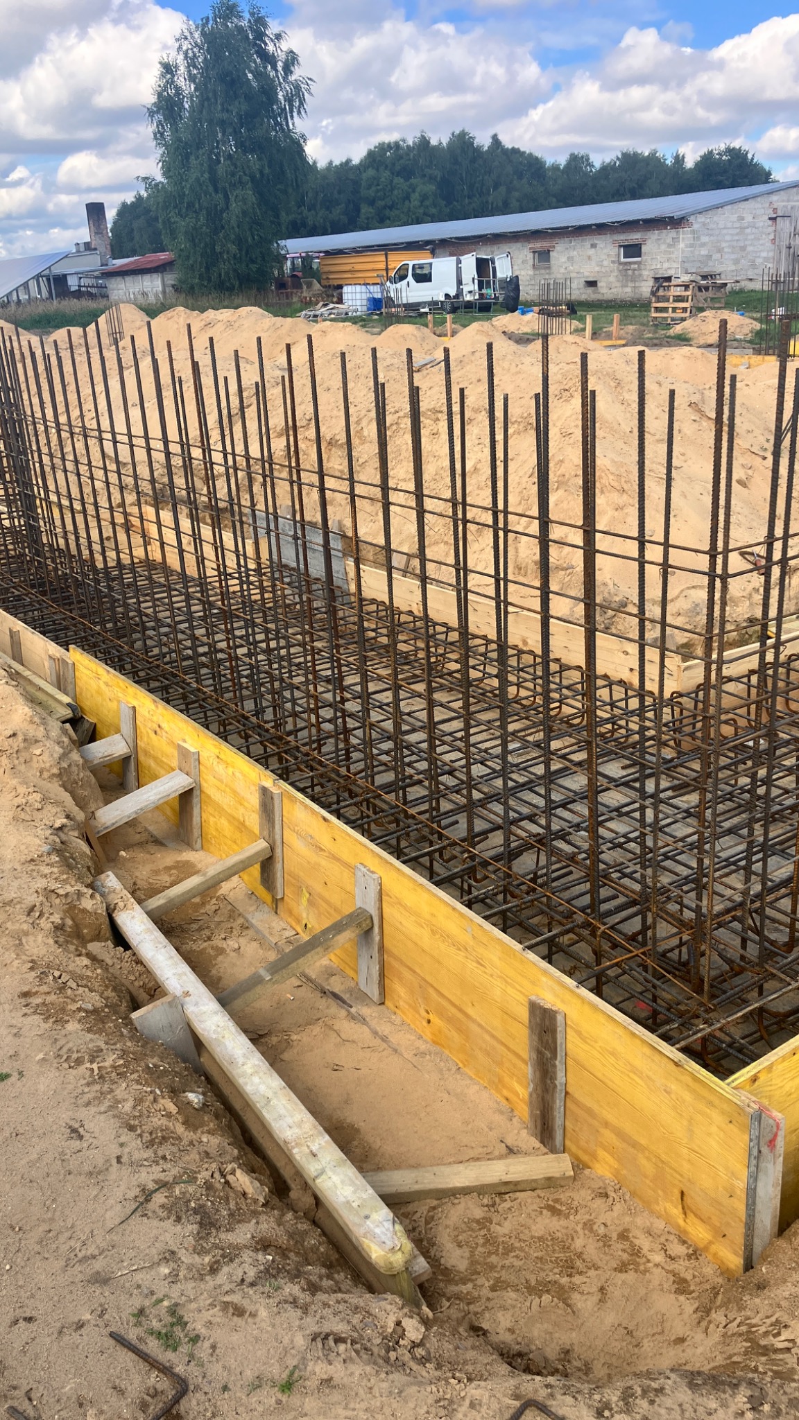 Zbrojenie fundamentów z drewnianym szalunkiem na placu budowy, widoczne pręty stalowe i przygotowane podłoże z piasku.