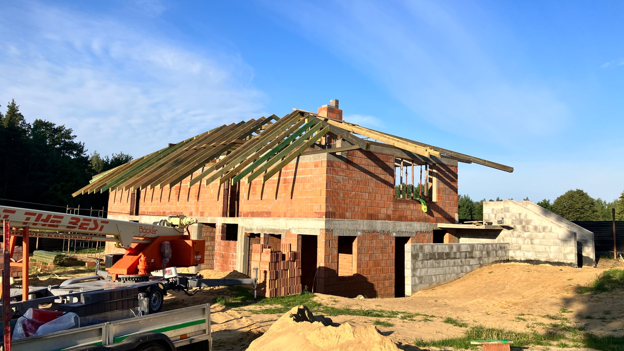 Wznoszona dwukondygnacyjna konstrukcja z czerwonej cegły z drewnianą konstrukcją dachu, widoczny dźwig budowlany i stosy cegieł, na tle błękitnego nieba i drzew.
