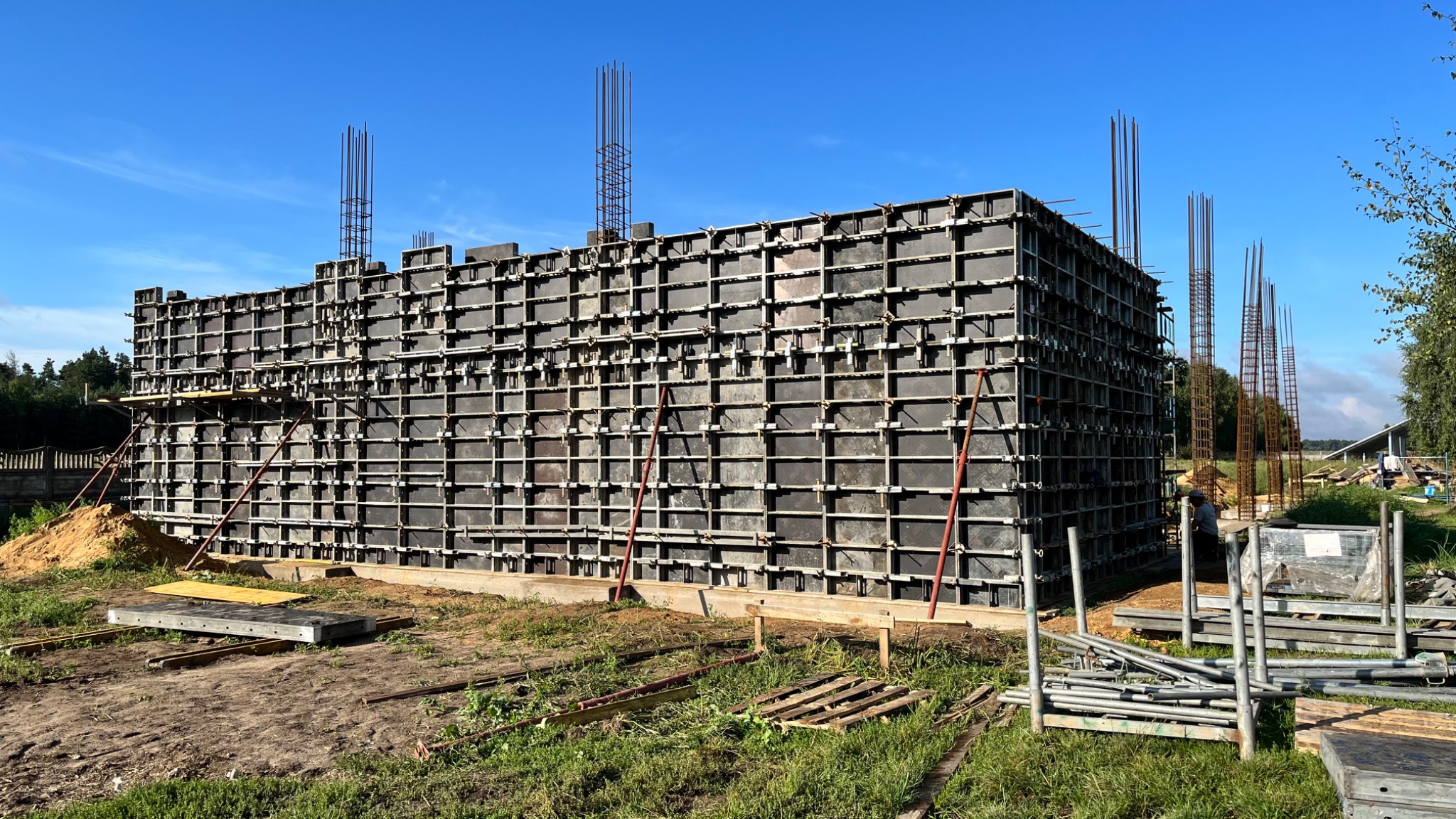 Szara szalunkowa konstrukcja z wystającymi prętami zbrojeniowymi na tle błękitnego nieba i zieleni, widoczne podpory i elementy konstrukcyjne.